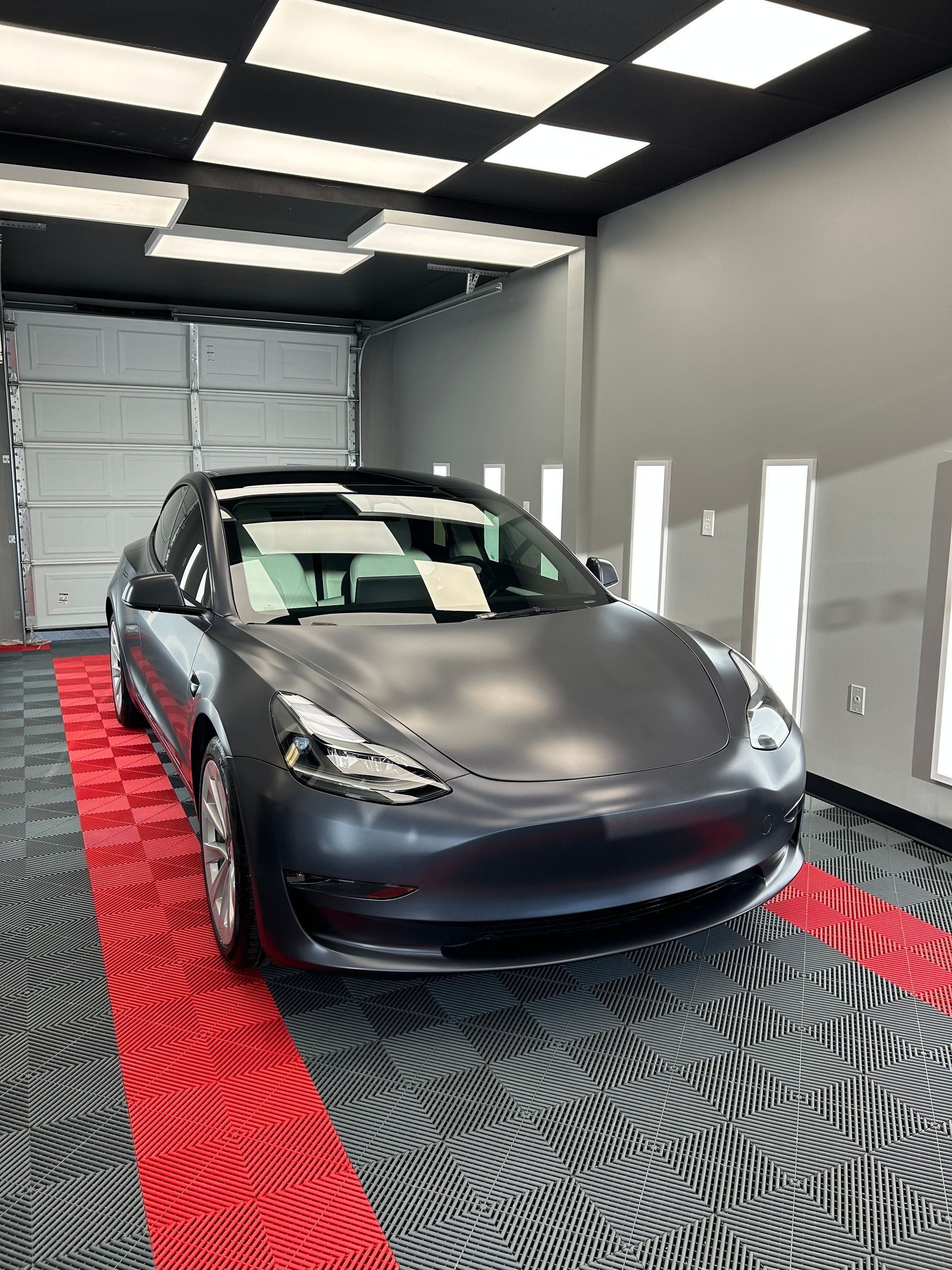 Dark gray Tesla car parked in a garage with red and black tiled floor and overhead lighting.