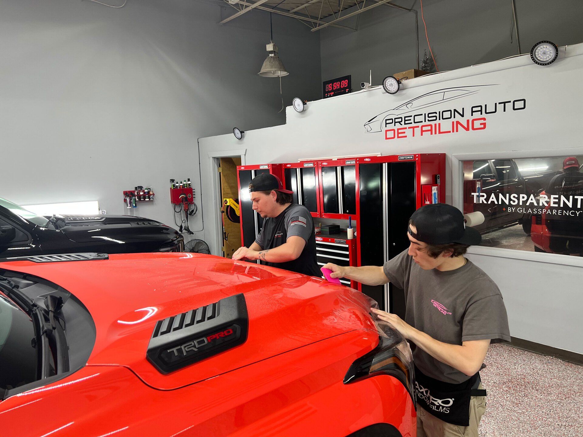 Two people applying film to a red car in a detailing shop.