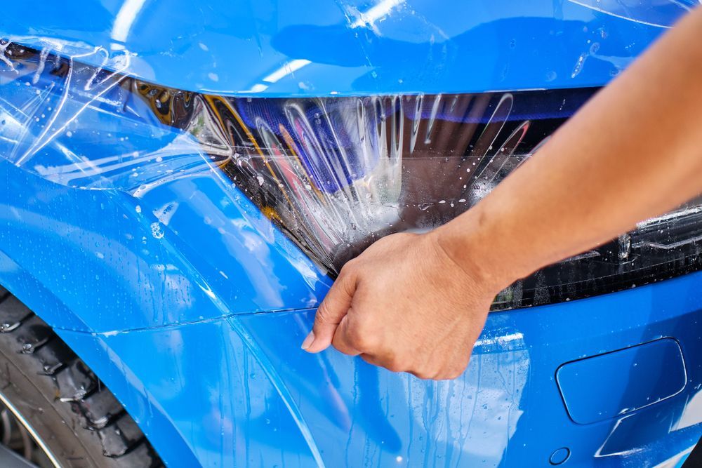Hand peeling clear protective film from a blue car bumper near the headlight