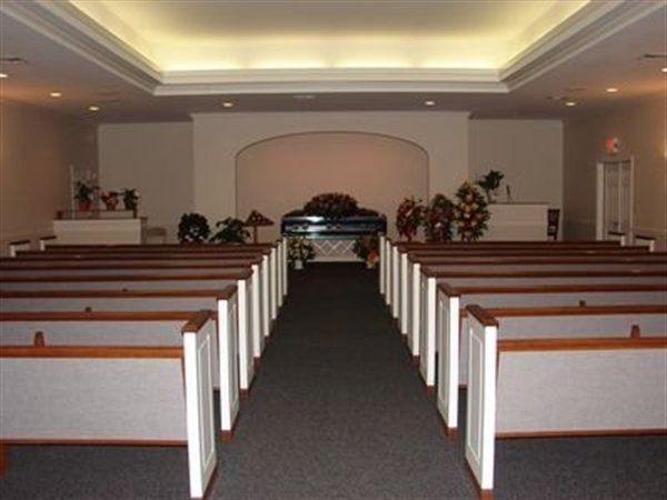 Funeral home interior with rows of empty pews facing a casket and floral arrangements.
