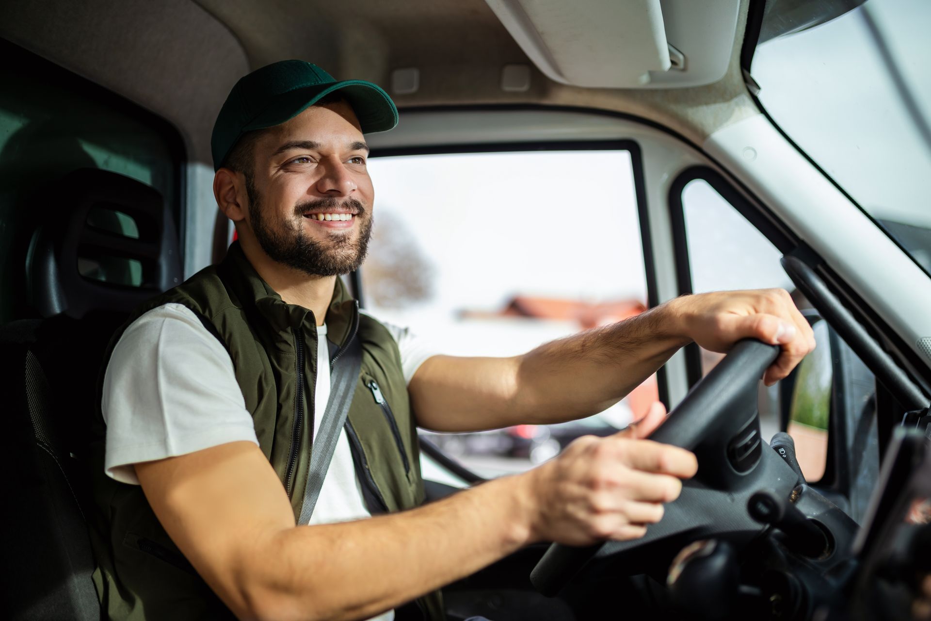A smiling delivery driver wearing a green cap and vest drives a vehicle while looking toward the road.