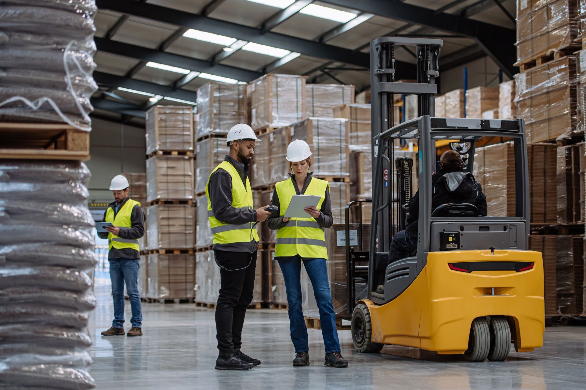 Warehouse workers in high-visibility vests consult paperwork near a forklift and stacked pallets of inventory.