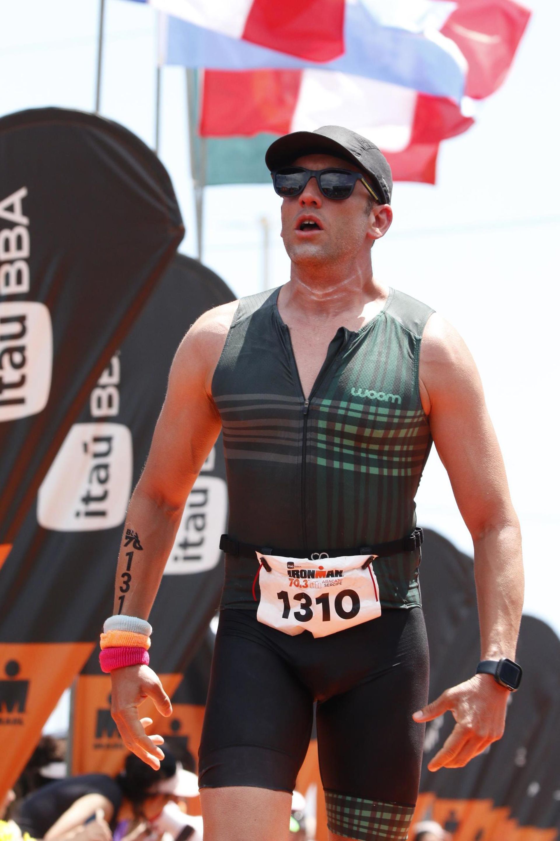 Triatleta na linha de chegada, vestindo roupa escura, número de peito 1310, óculos de sol, boné, com bandeiras ao fundo.