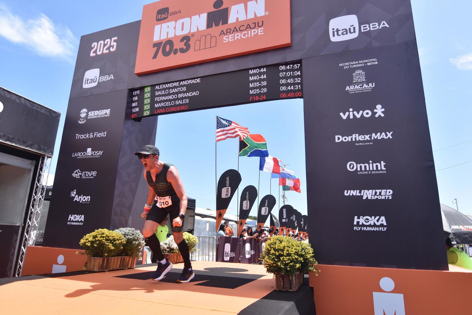 Triatleta cruzando a linha de chegada do Ironman 70.3, com os braços erguidos, sob uma faixa com bandeiras em Aracaju