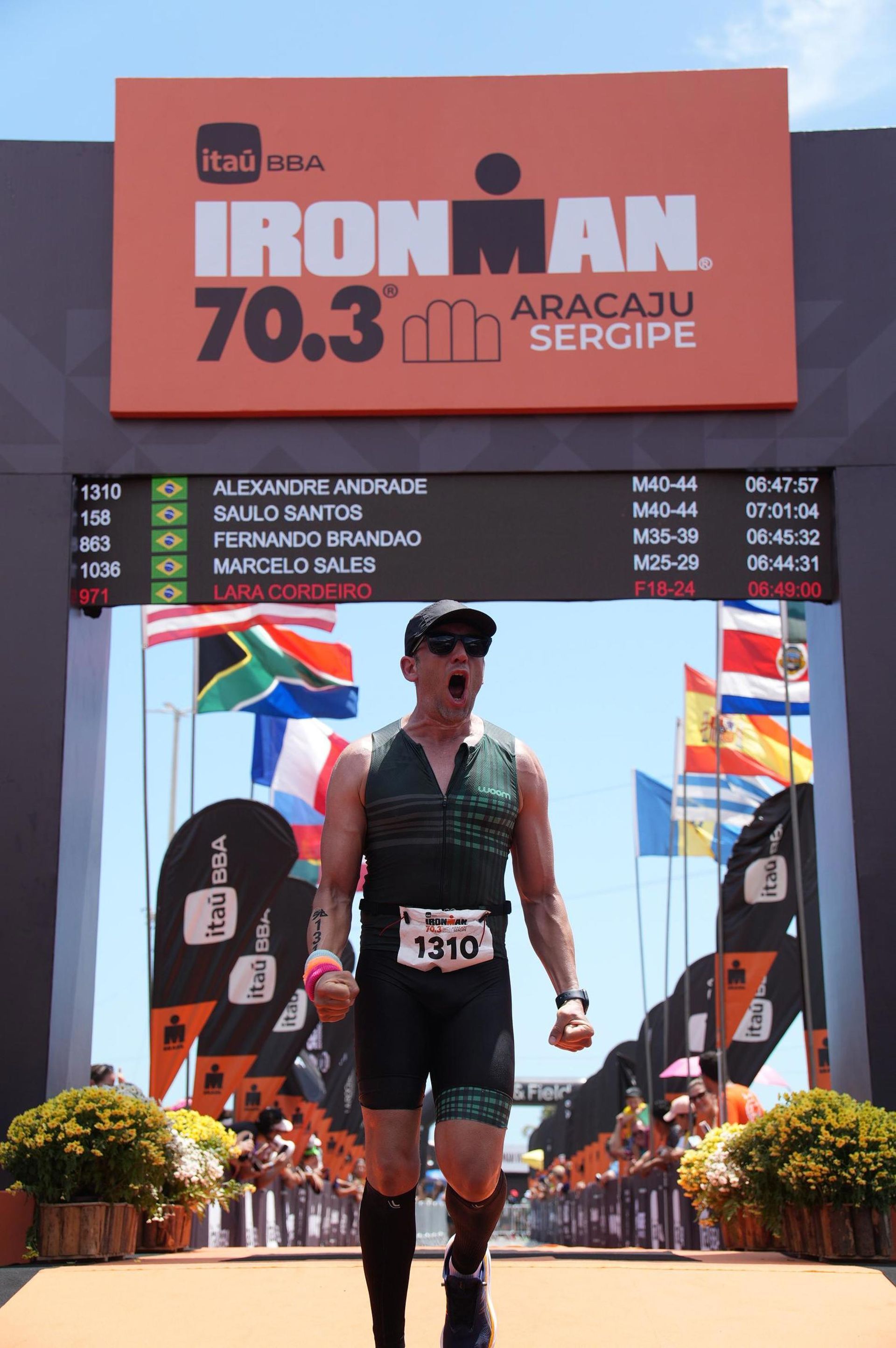 Triatleta cruza a linha de chegada do Ironman 70.3 em Aracaju, Sergipe, comemorando com os punhos cerrados.