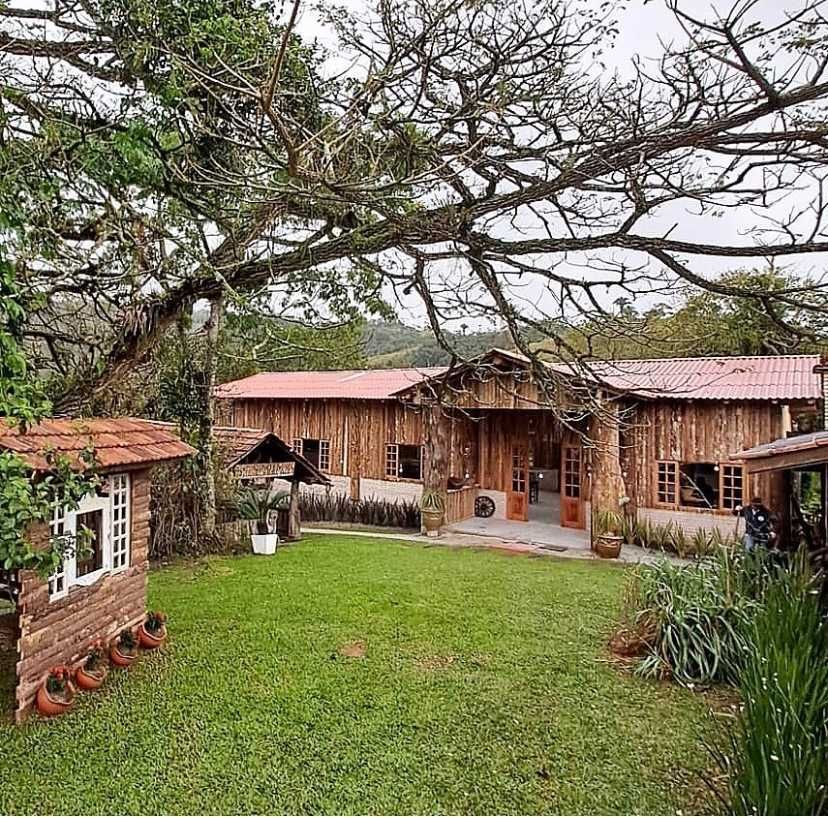 Cabana de madeira com telhado vermelho, casa pequena e gramado verde sob uma grande árvore.