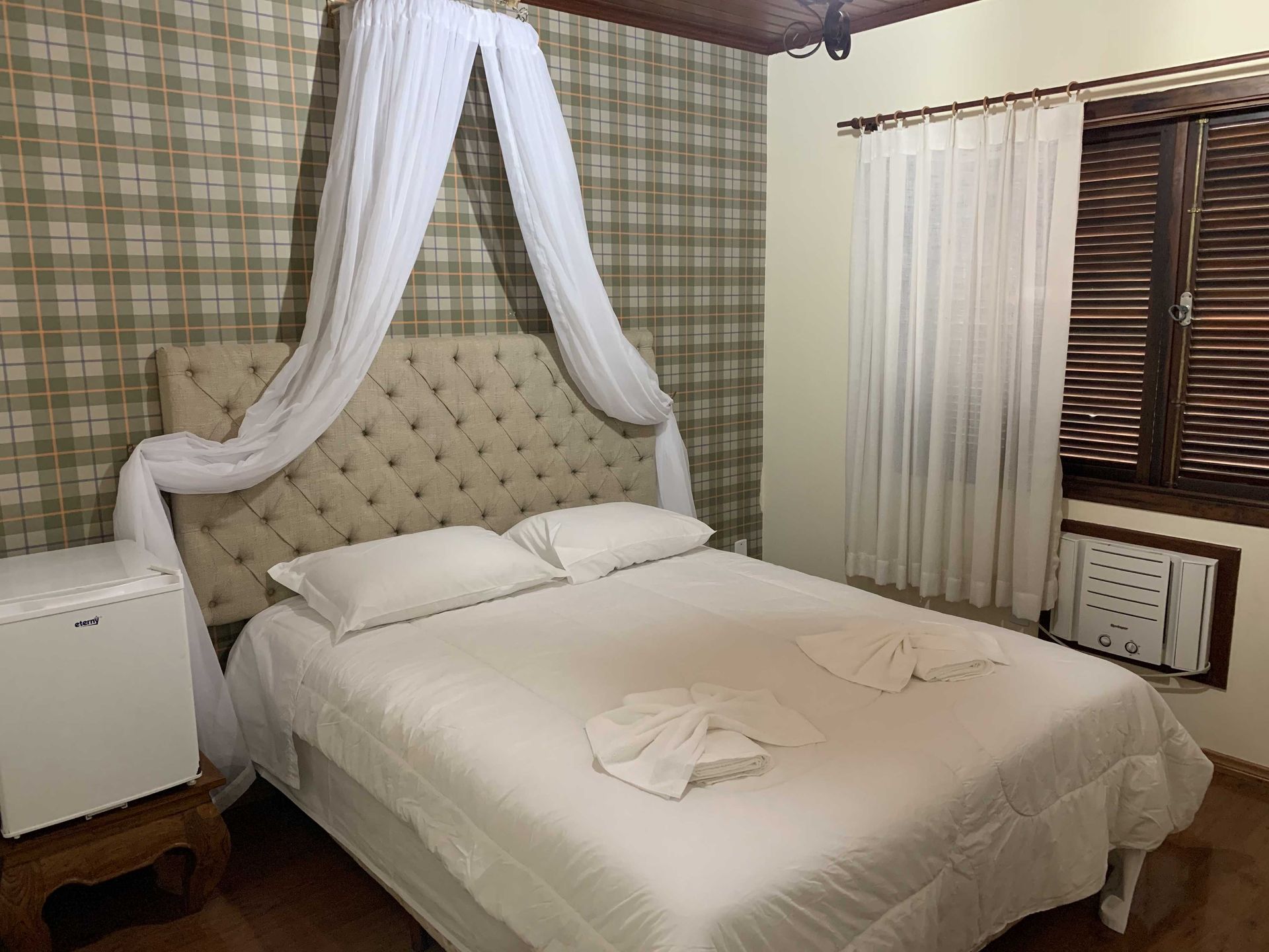 Quarto com cama de casal, cabeceira estofada, dossel transparente, papel de parede estampado e janela fechada com ar condicionado.