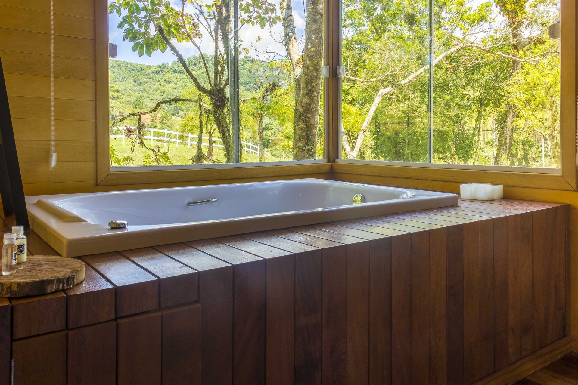 Banheiro com painéis de madeira, banheira retangular e grande janela com vista para uma paisagem verdejante.