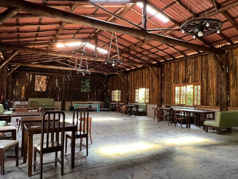 Interior rústico de madeira com mesas, cadeiras e bancos. A luz entra pelas janelas.