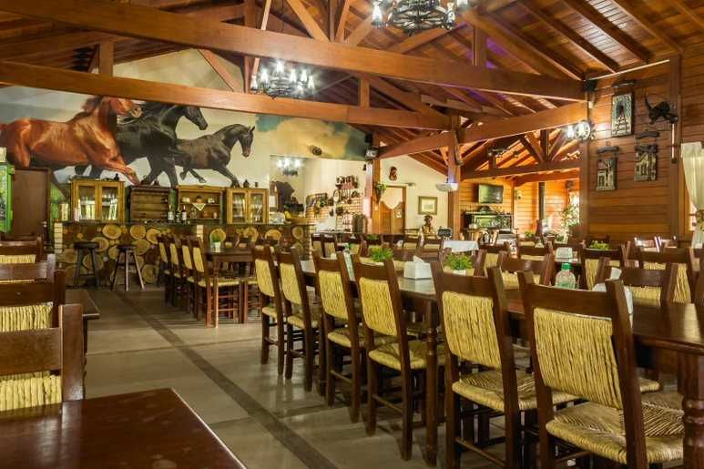Interior de restaurante com mesas e cadeiras de madeira, mural de cavalos e lustres decorativos.