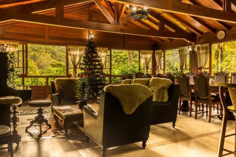 Sala aconchegante com teto de madeira, árvore de Natal, poltronas, mesa de jantar e grandes janelas com vista para uma vegetação exuberante.