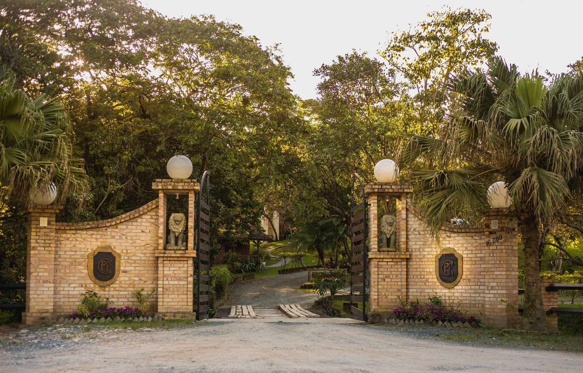 Portão de entrada de tijolos com luzes globulares brancas, que dá acesso a um caminho ladeado por árvores.