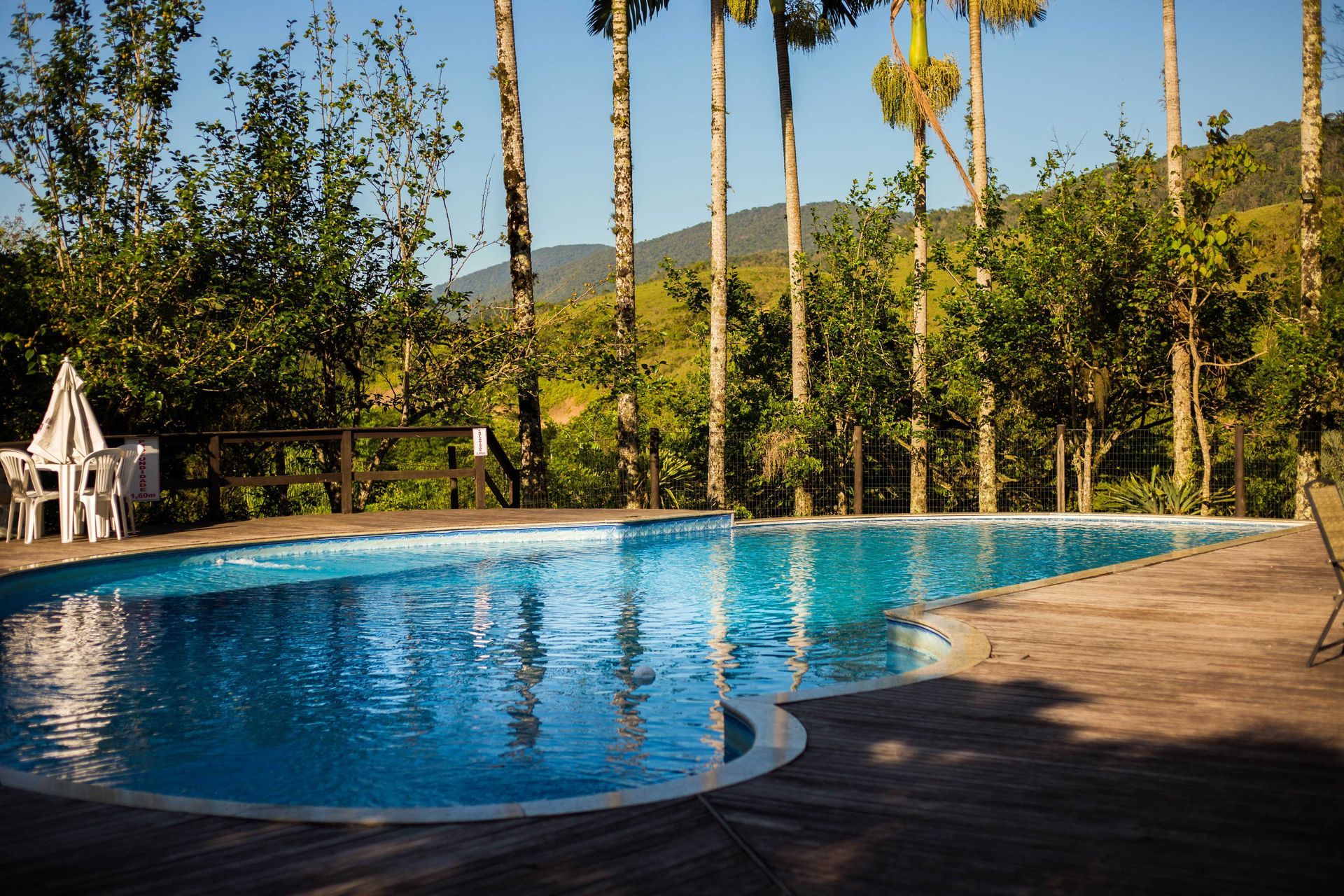 Piscina rodeada por árvores, deck de madeira e vista para as montanhas em um dia ensolarado.