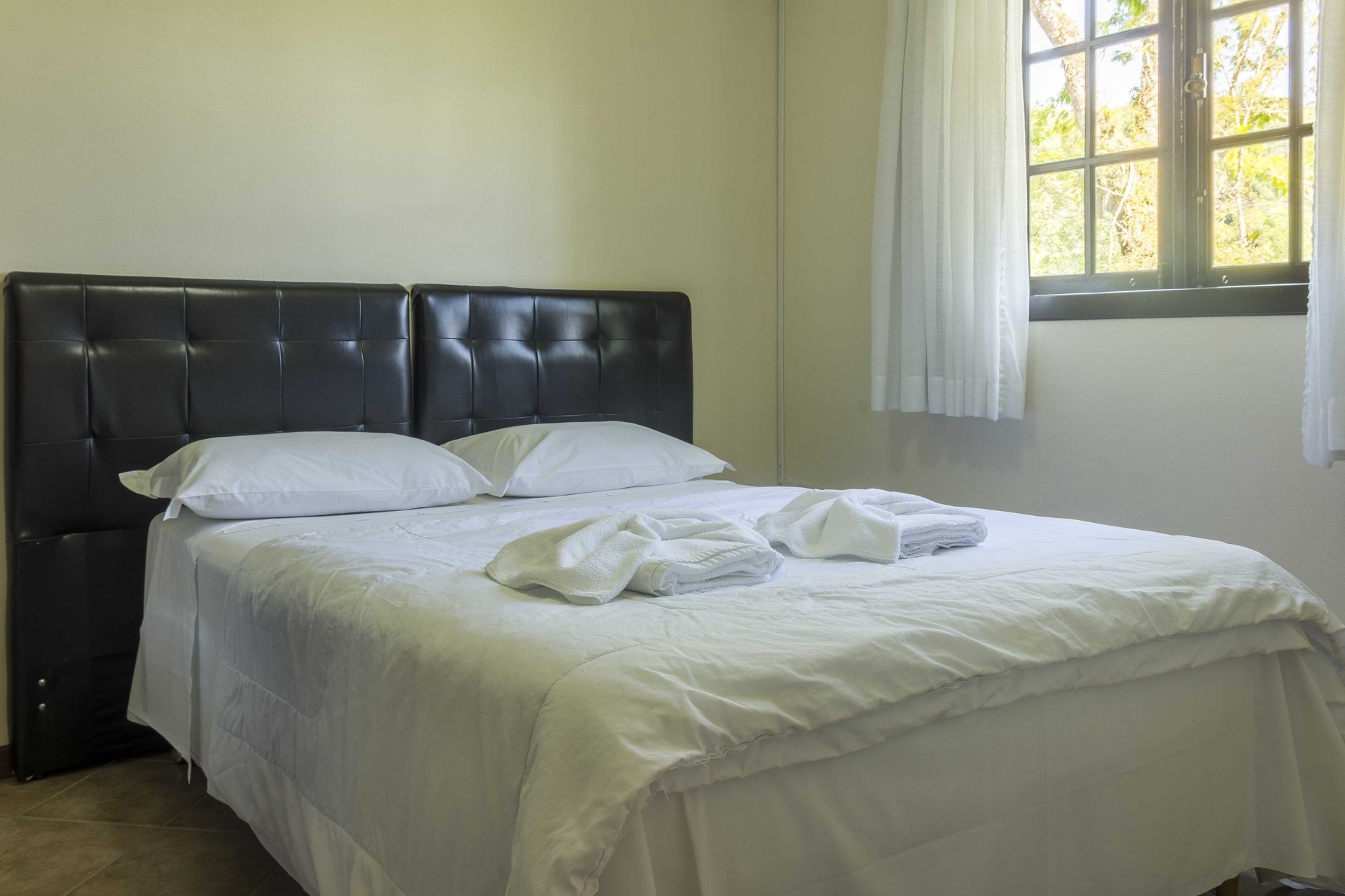 Uma cama com lençóis brancos, dois travesseiros e toalhas dobradas. Há uma cabeceira preta e uma janela à direita.