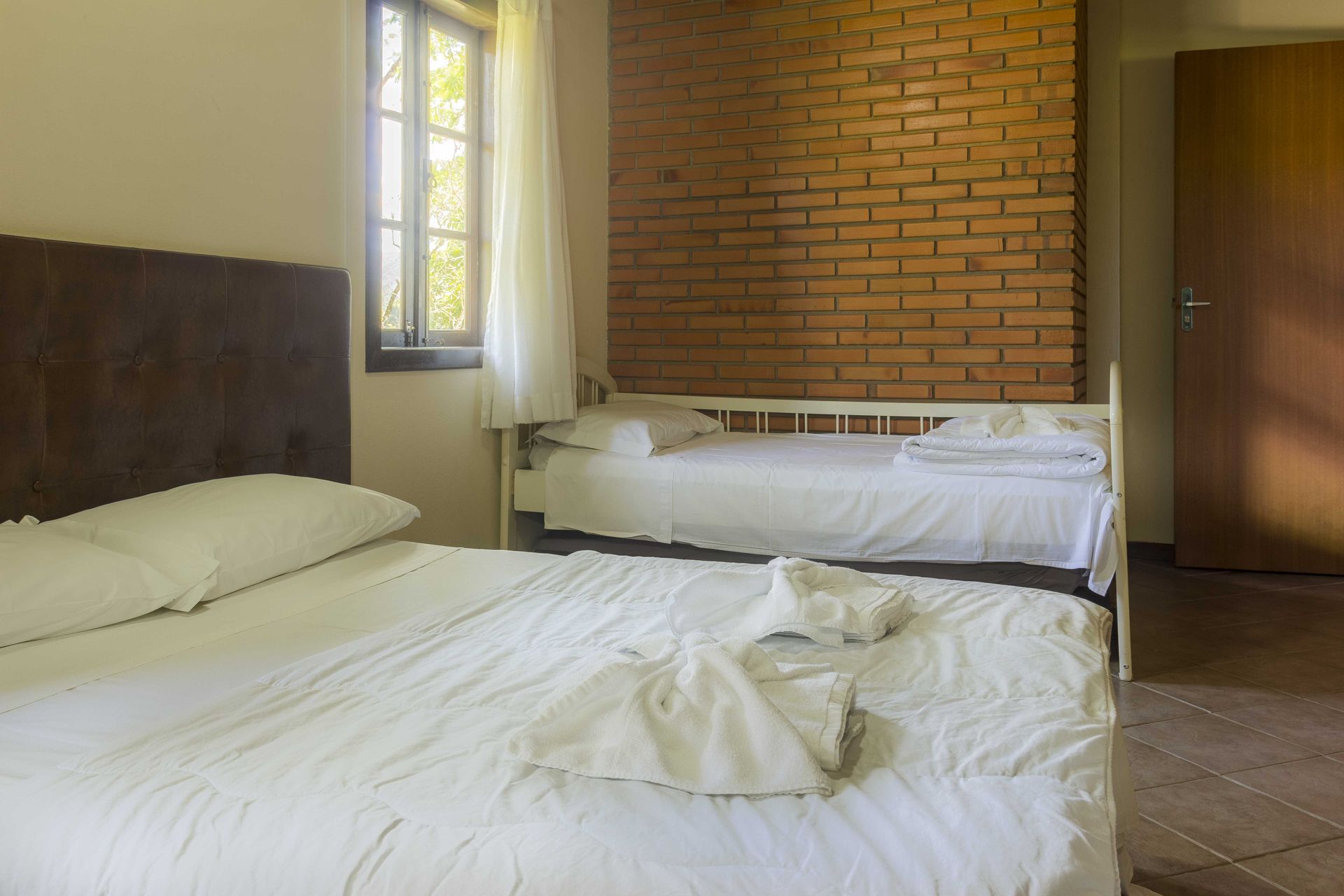 Quarto com duas camas, uma grande e uma menor, encostado a uma parede de tijolos e com janela.