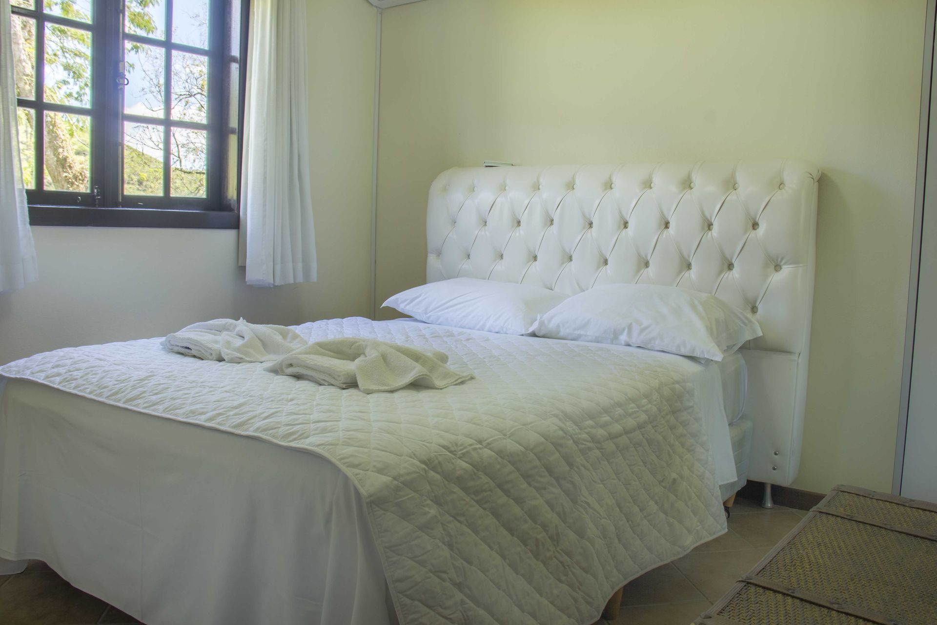 Cama branca em um quarto de cores claras com janela, lençóis brancos e cabeceira.