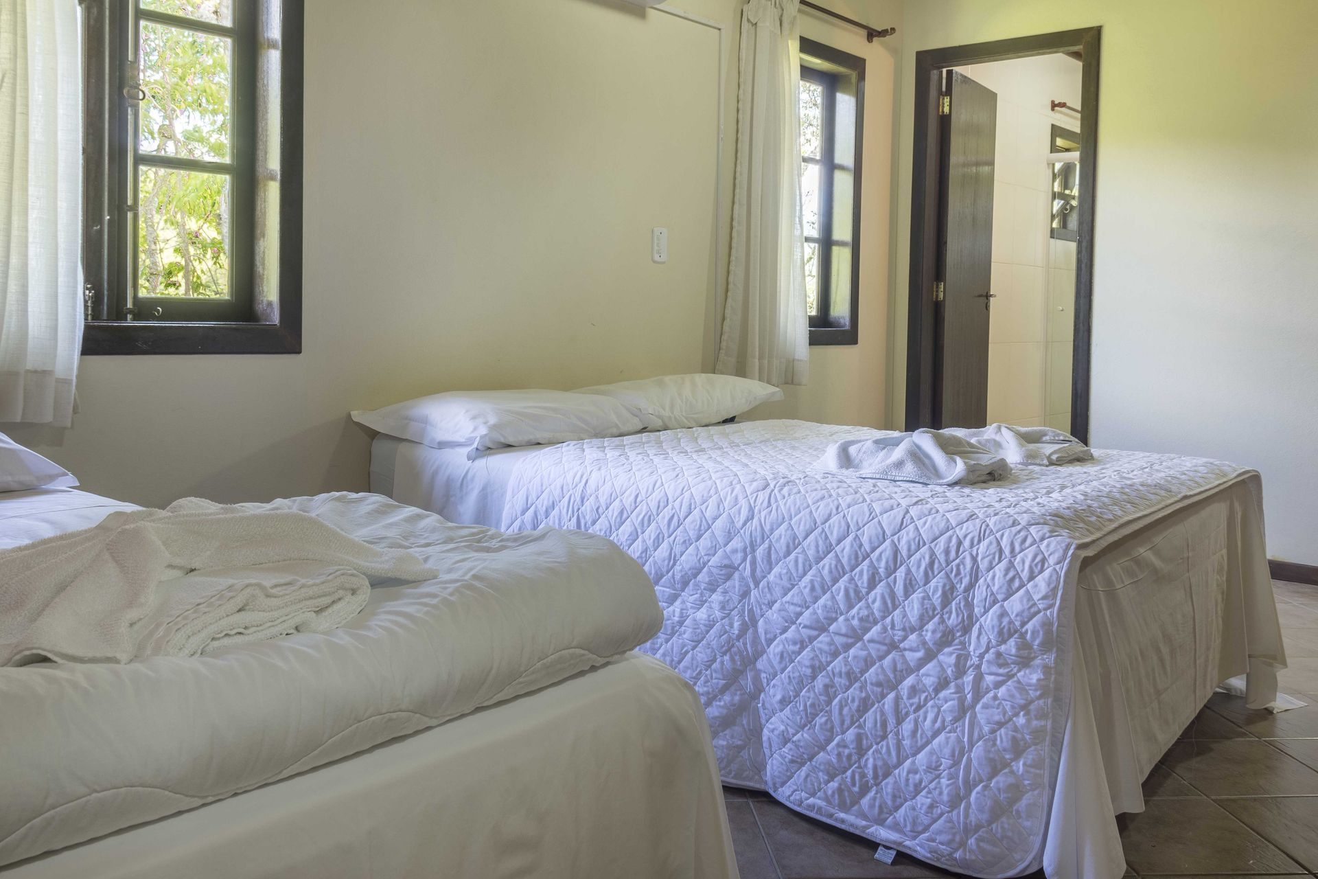 Duas camas em um quarto com roupa de cama branca, perto de uma janela e porta aberta.