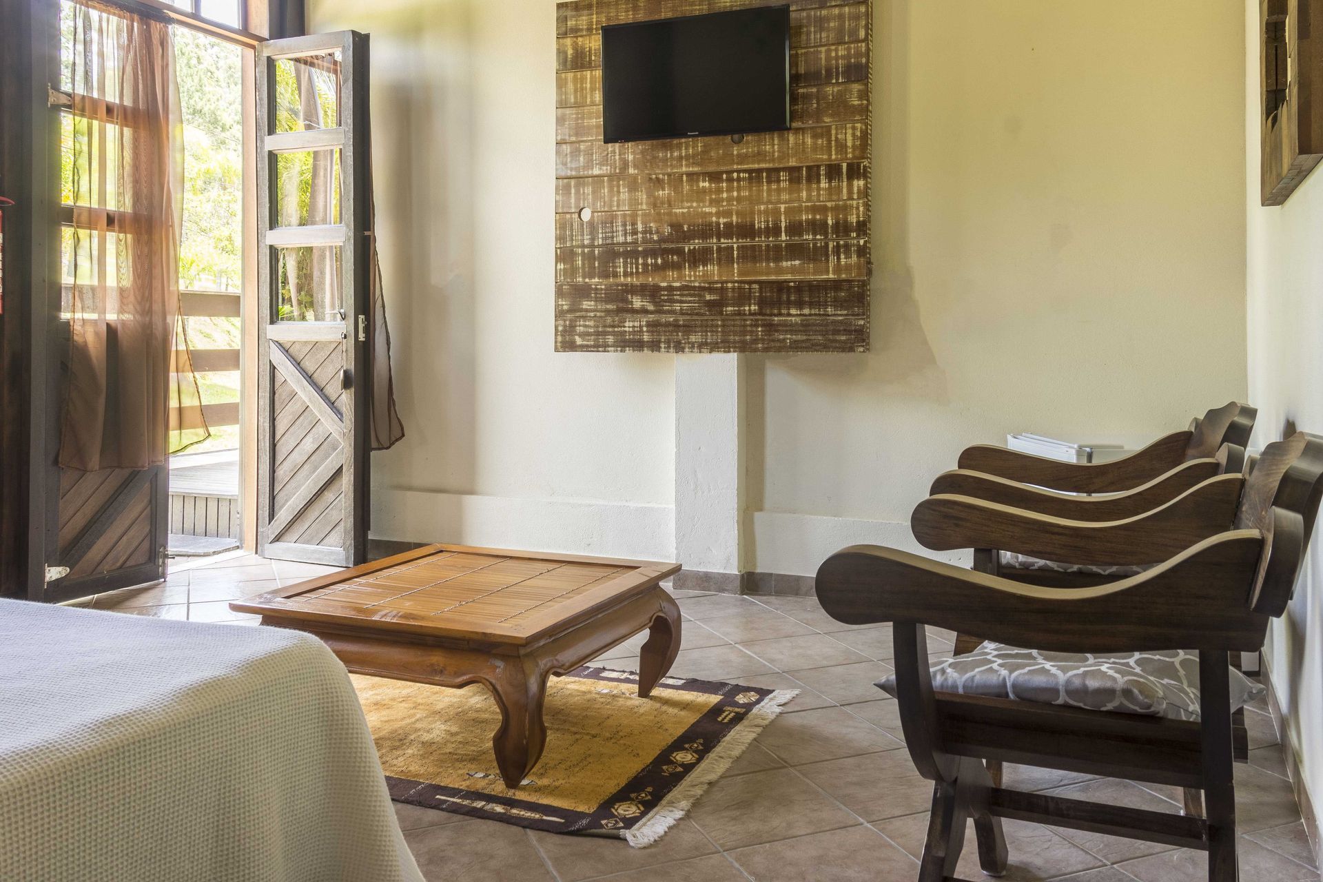 Sala de estar com porta de madeira aberta, TV, cadeiras de madeira e mesa de centro sobre um tapete.