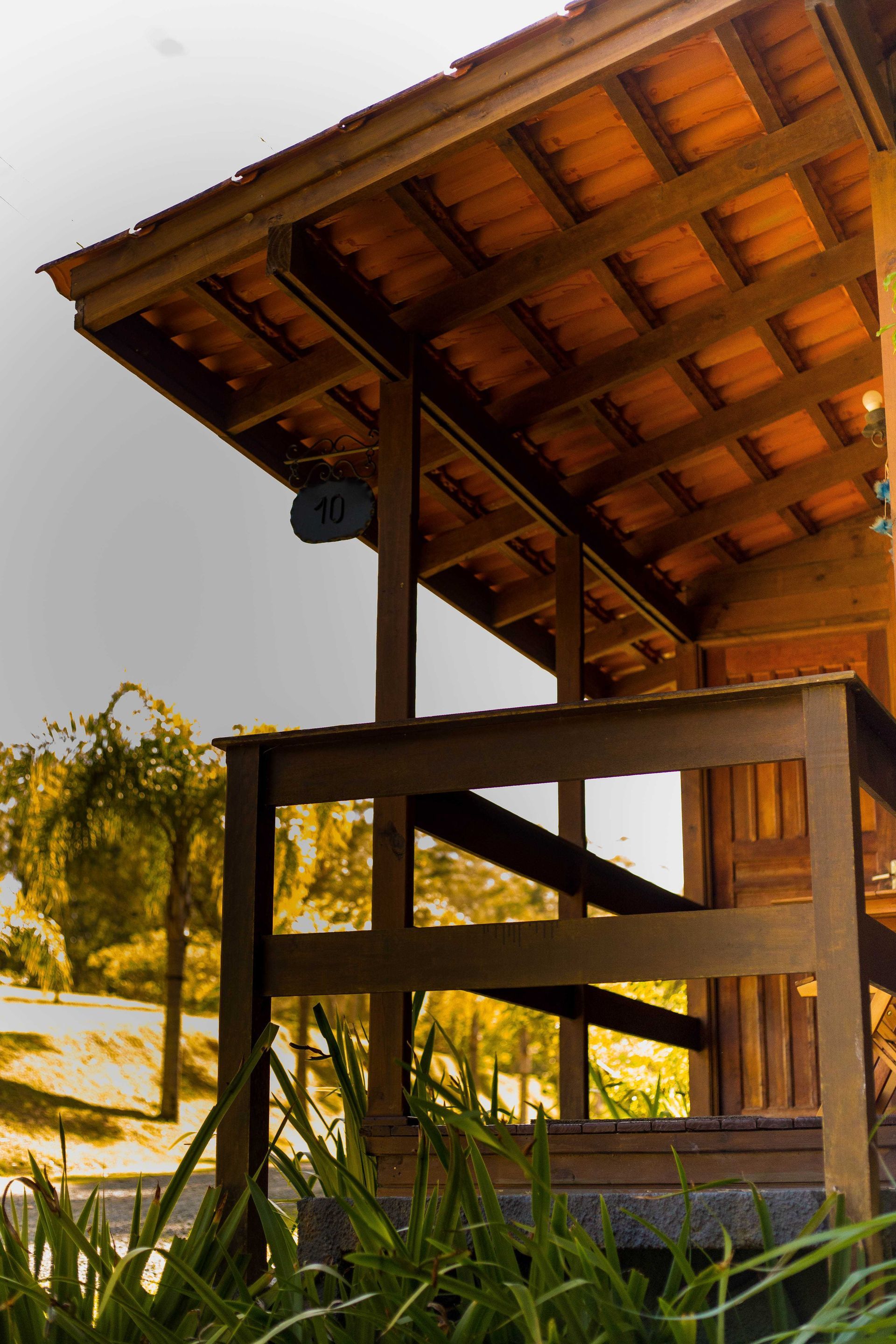 Varanda de madeira com telhado de telha, corrimão e folhagem em um ambiente externo de tons quentes.