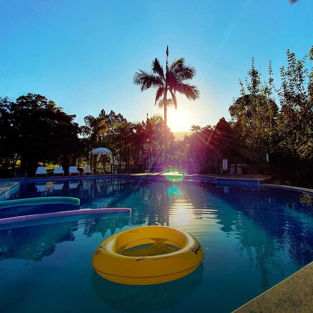 Piscina com uma boia amarela, água azul e uma palmeira em silhueta contra o sol ao pôr do sol.