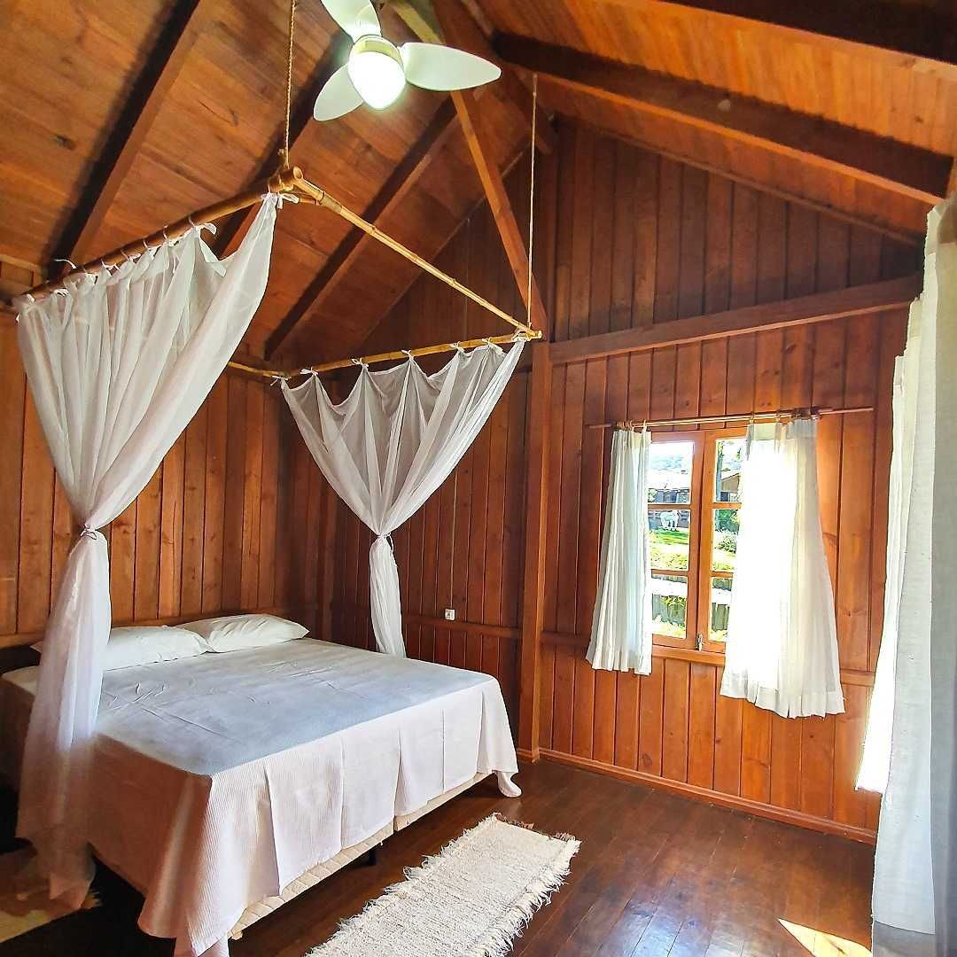 Quarto aconchegante de madeira com cama de dossel, janela com cortinas e tapete branco.