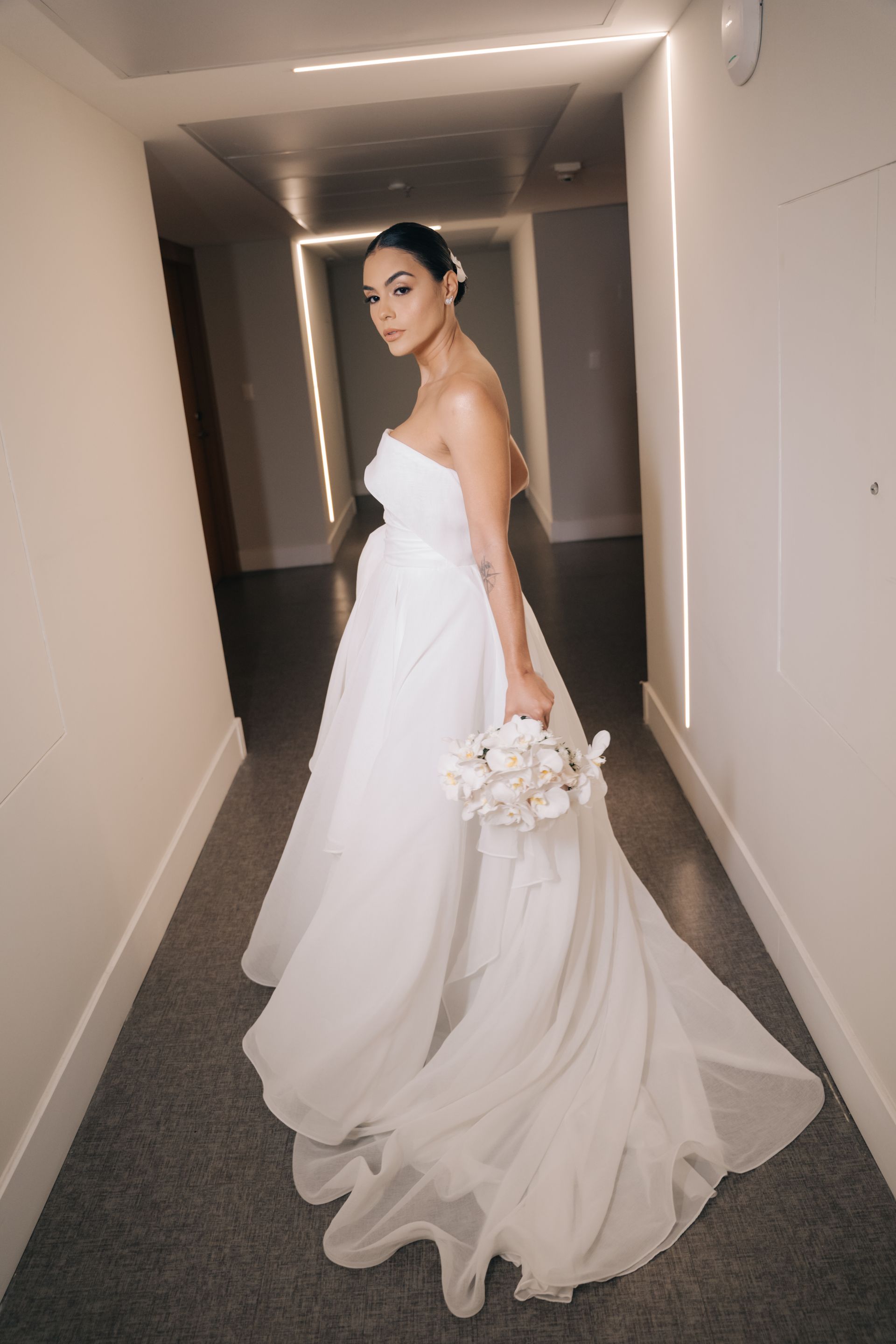 Uma mulher vestida de noiva está parada em um corredor segurando um buquê de flores.