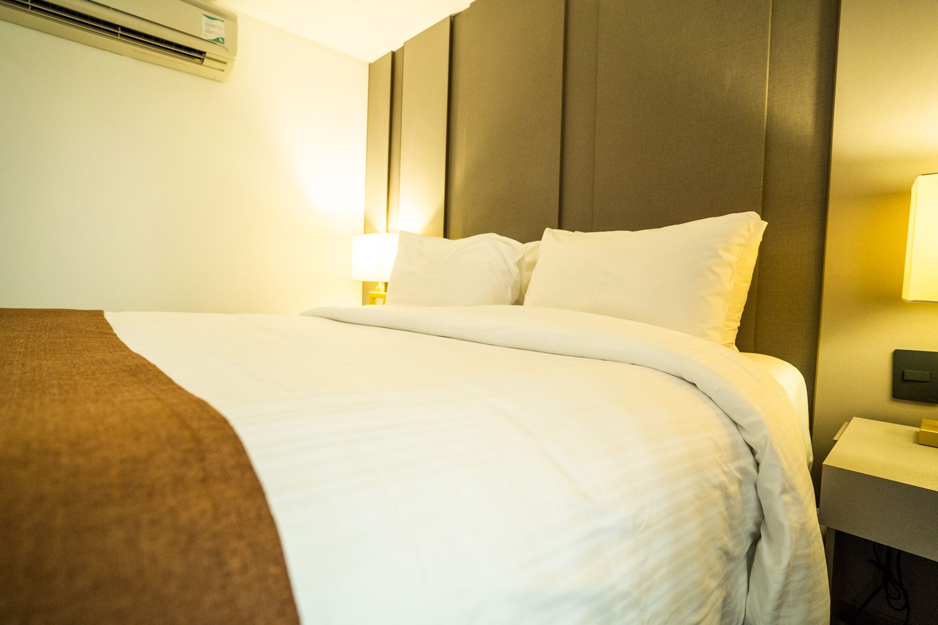 Um quarto de hotel com cama, mesa de cabeceira, abajur e ar condicionado.