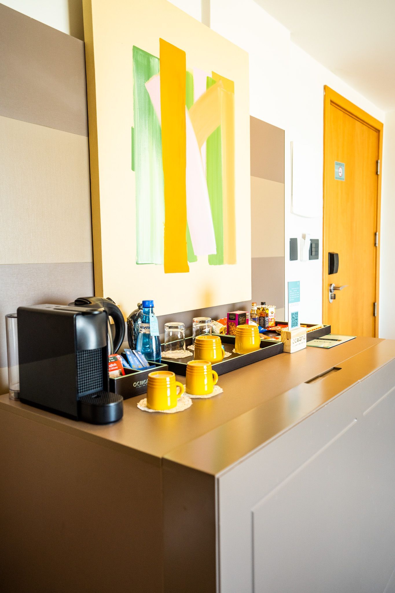Um quarto de hotel com uma cafeteira, xícaras e uma pintura na parede.