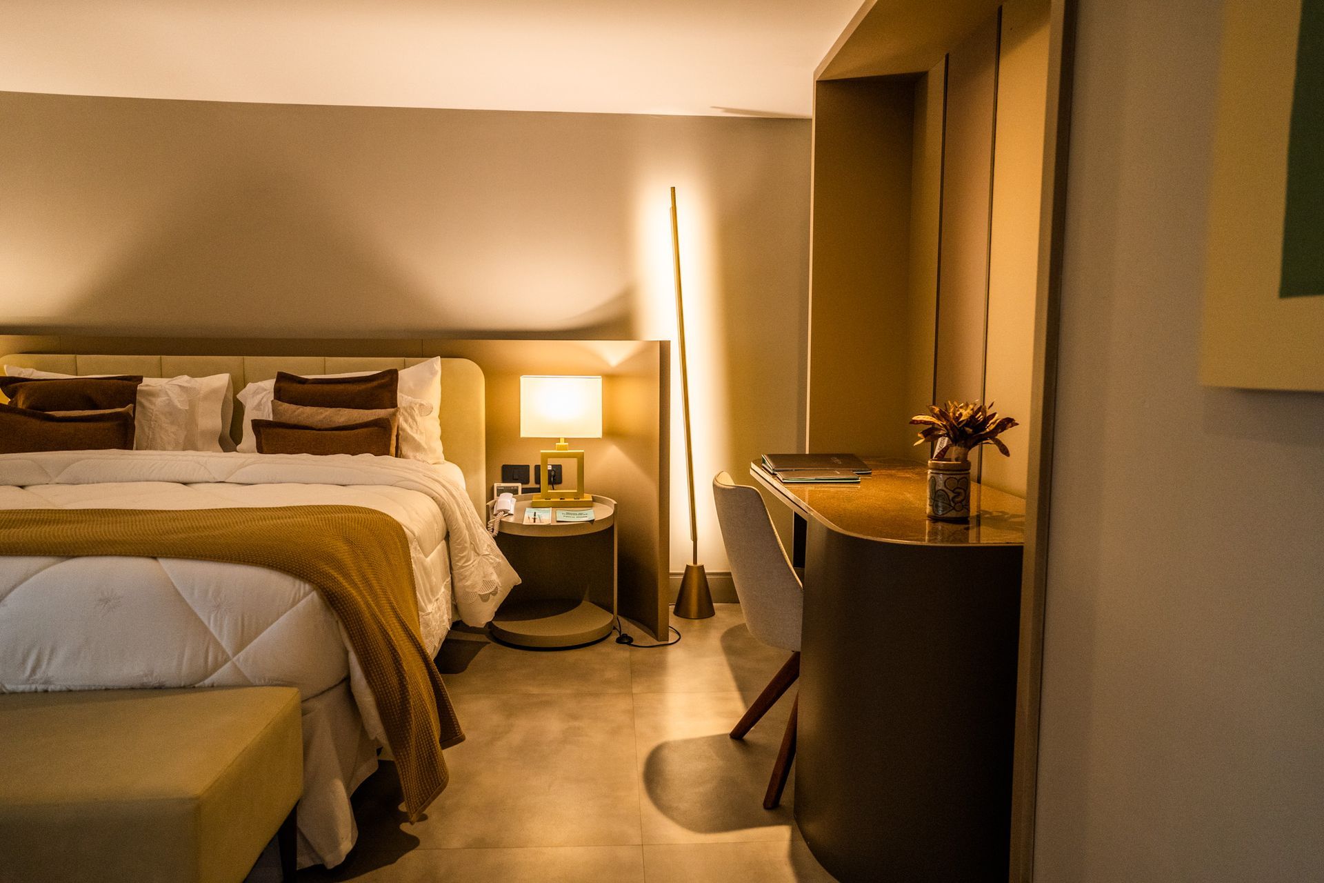 Um quarto de hotel com uma cama king size, escrivaninha, mesa de cabeceira e luminária.