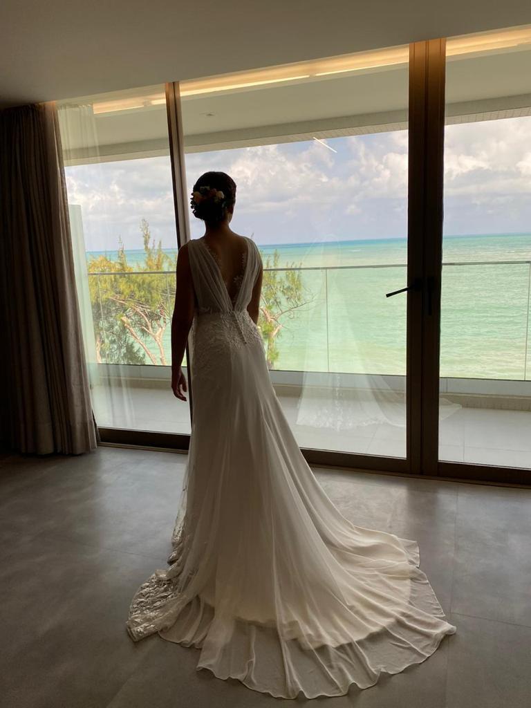 Uma mulher vestida de noiva está parada em frente a uma janela olhando para o oceano.