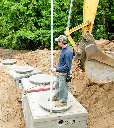 A worker stands on a concrete septic tank lid at an outdoor construction site, holding a measuring pole near an excavator.