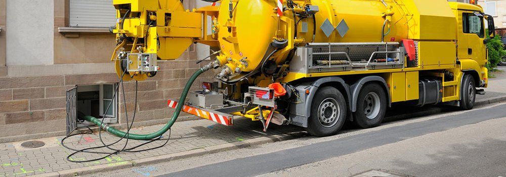 A yellow vacuum truck parked on a street, using a green hose to suction material from a building basement access point.