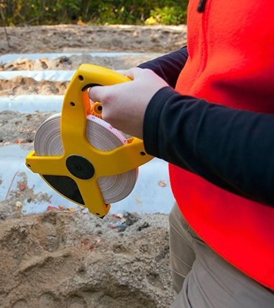 A person in an orange vest holds a yellow surveying tape measure over a sandy area with rows of plastic sheeting.