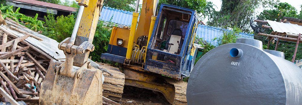 A yellow excavator sits in an outdoor area next to a large, grey, cylindrical water tank.