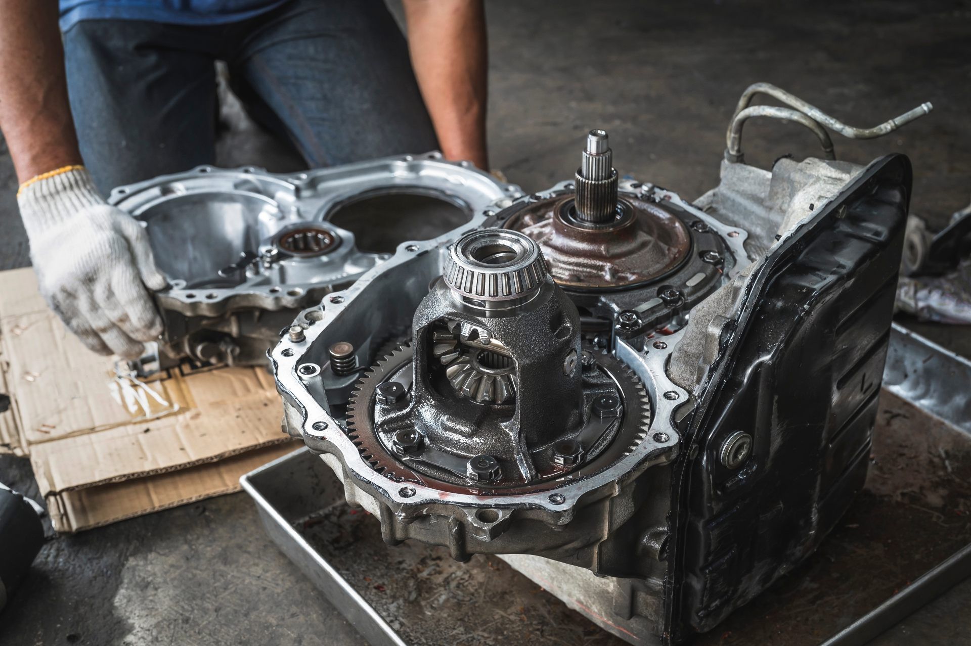 A mechanic in gloves disassembles an automotive transmission on a metal tray in a workshop.