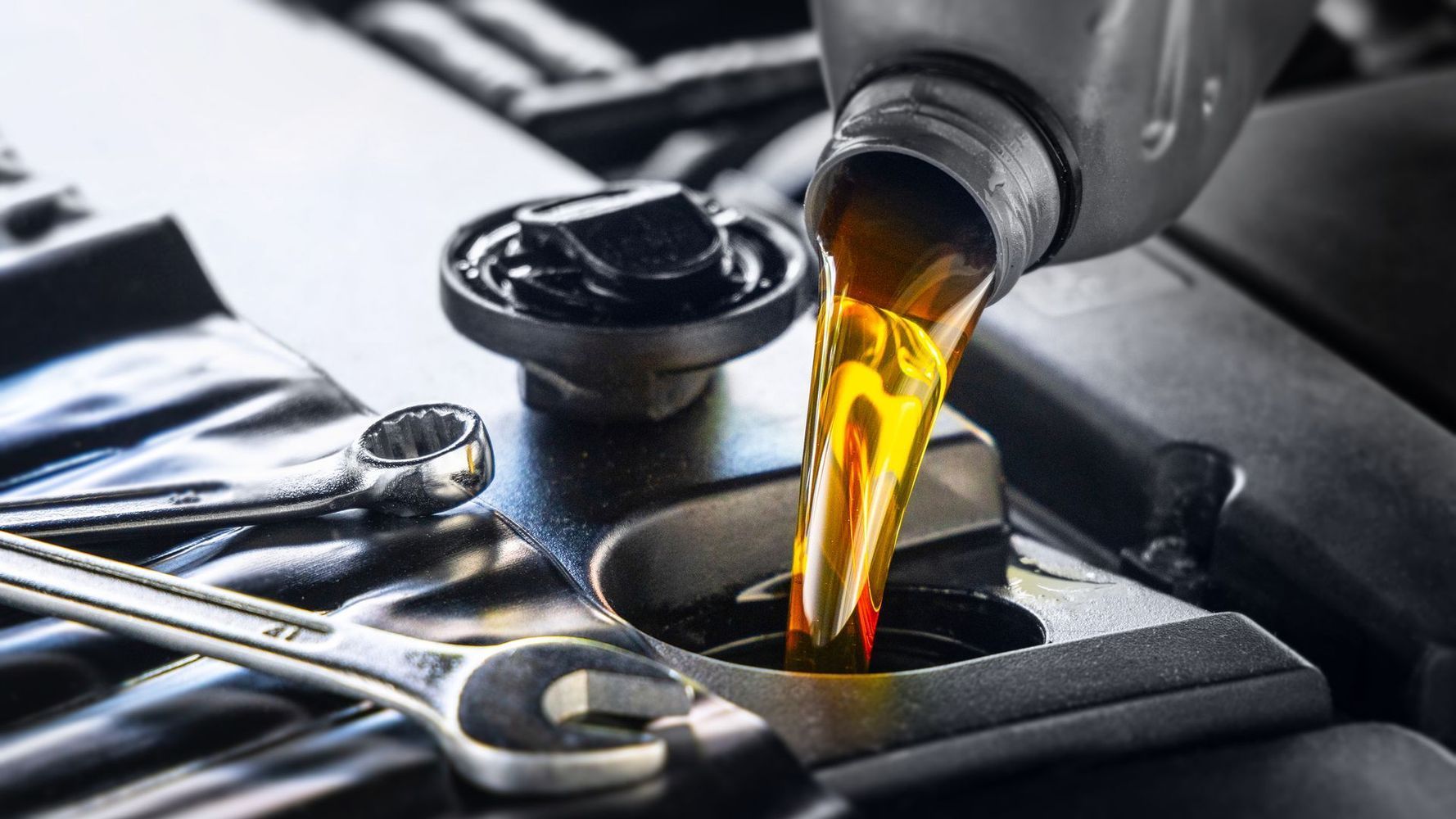 New engine oil being poured into a car engine, with two metal wrenches resting nearby on the engine cover.