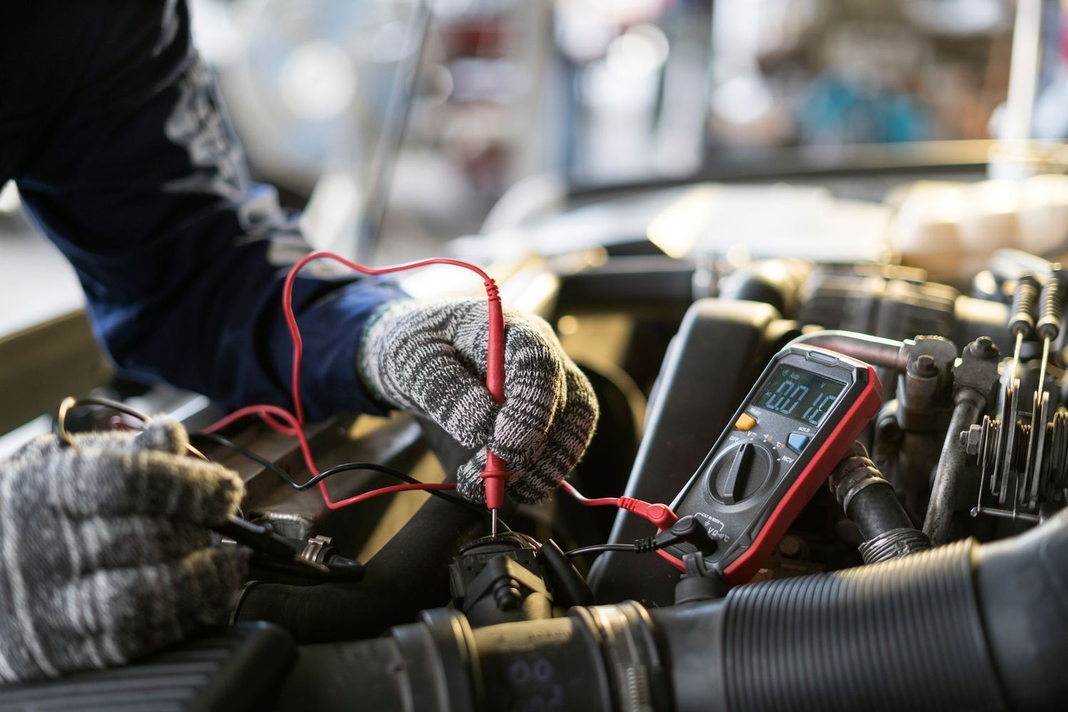 A mechanic wearing grey gloves uses a multimeter to test electrical components inside a vehicle engine bay.