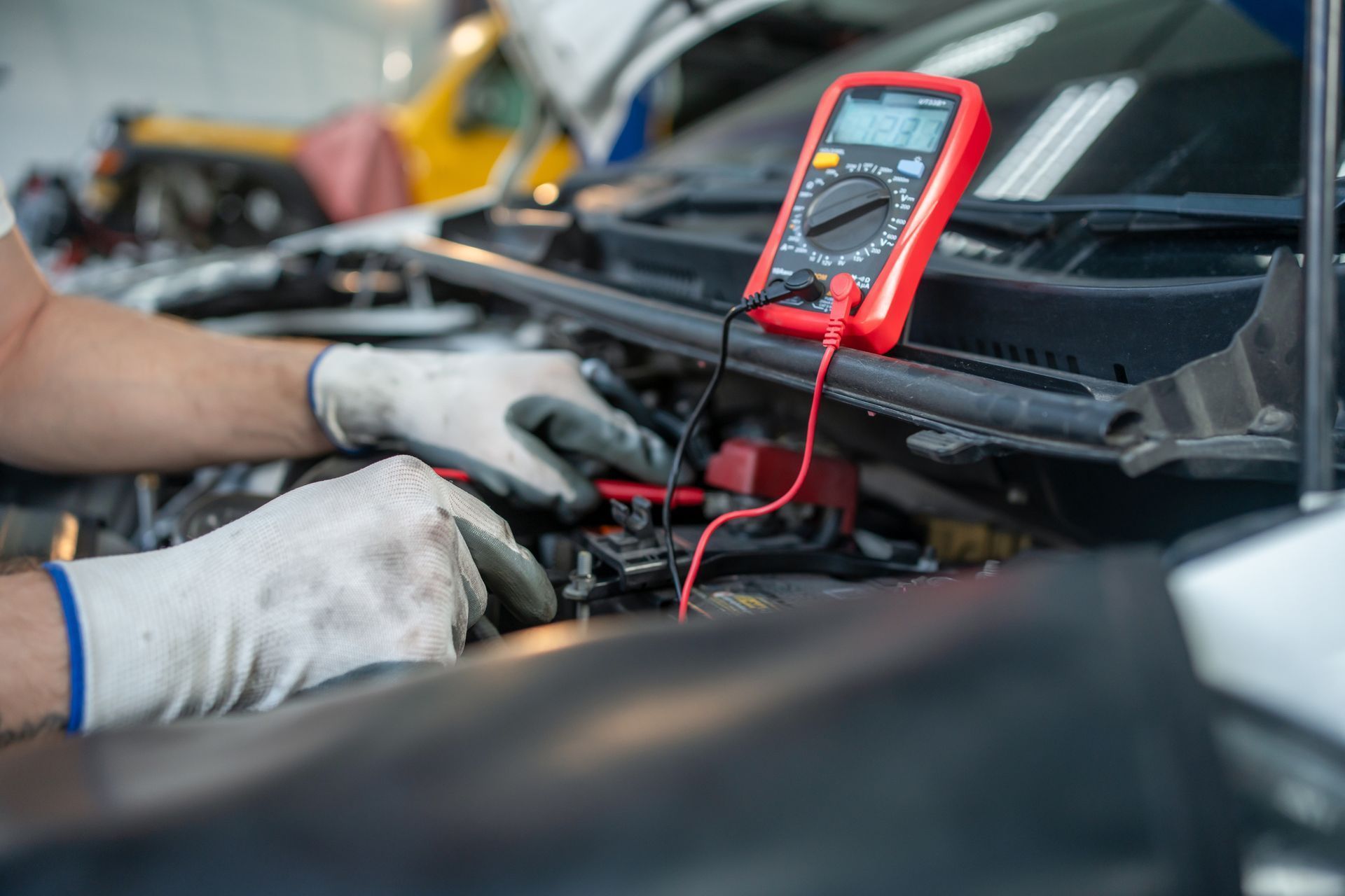 A mechanic wearing work gloves uses a red digital multimeter to test a car battery under the hood of a vehicle.