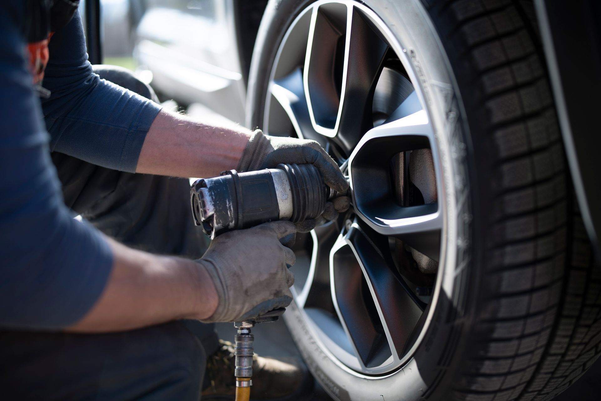 A mechanic in work gloves uses a pneumatic impact wrench to tighten a lug nut on a car wheel.