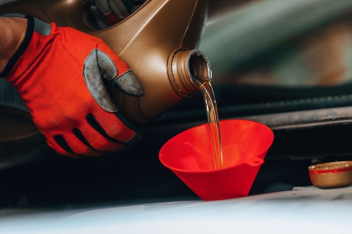 A person wearing bright orange work gloves pours golden motor oil from a plastic jug through a red funnel.
