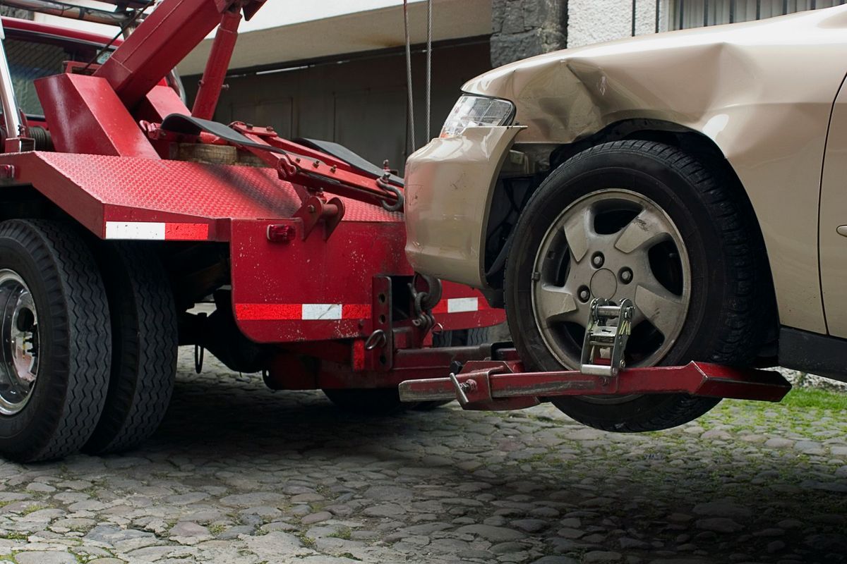 A red tow truck hook lifting the front tire of a beige car with a dented fender on a cobblestone surface.