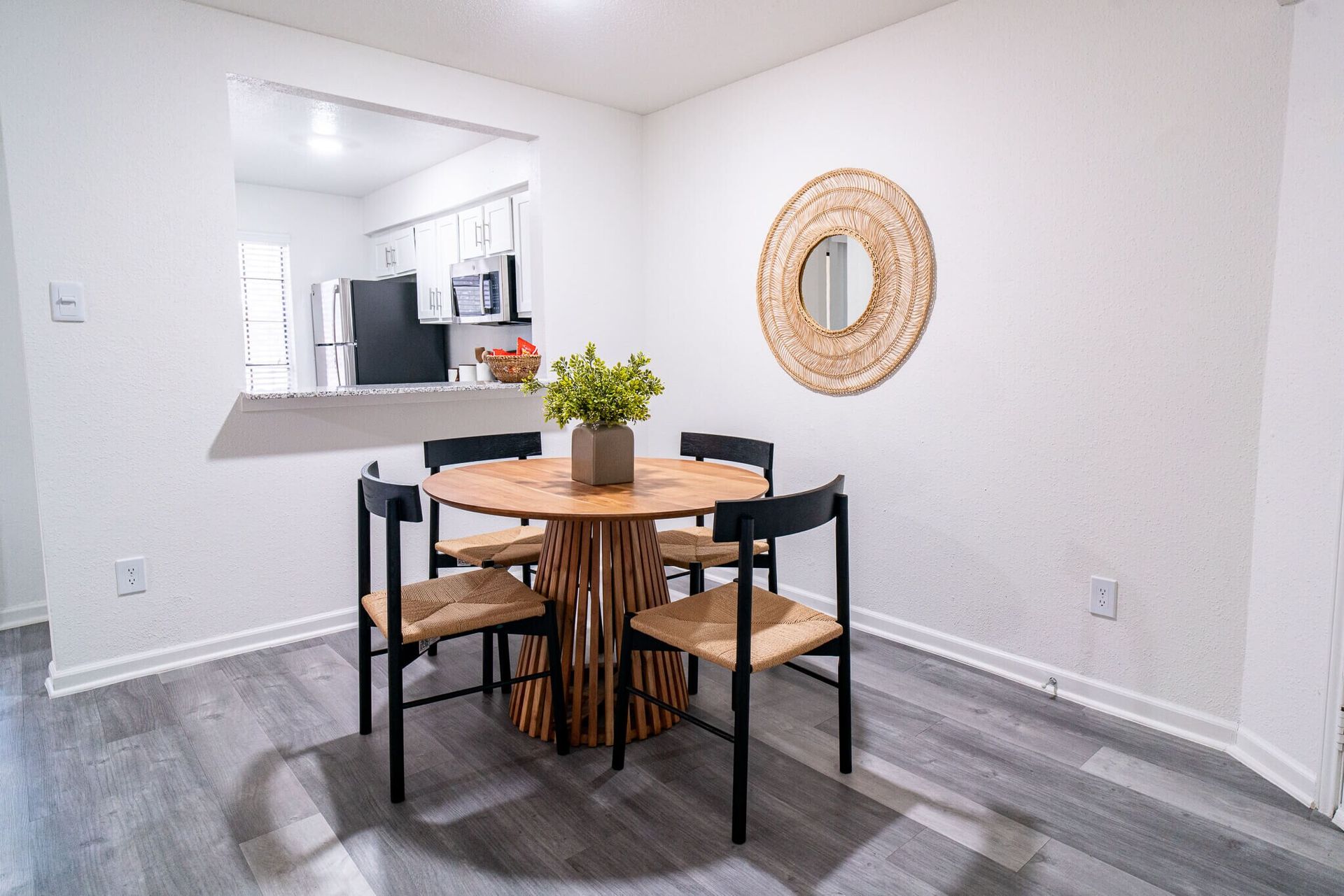 A dining area with a round wooden table, four black chairs, a small green plant, and a decorative mirror on the wall.