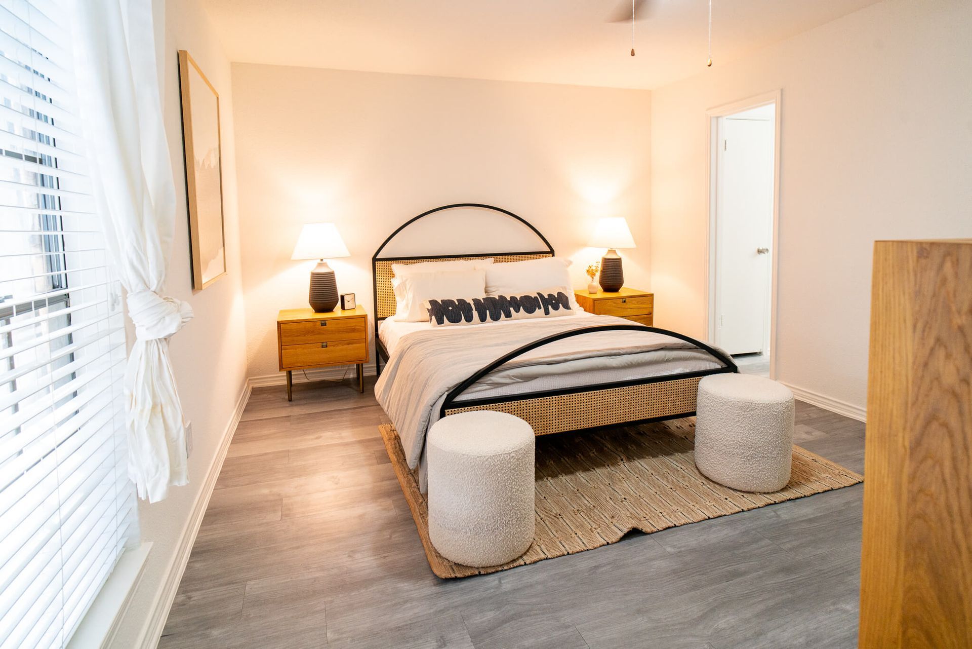 Bright bedroom with a queen bed, two wooden nightstands, and two round ottomans.