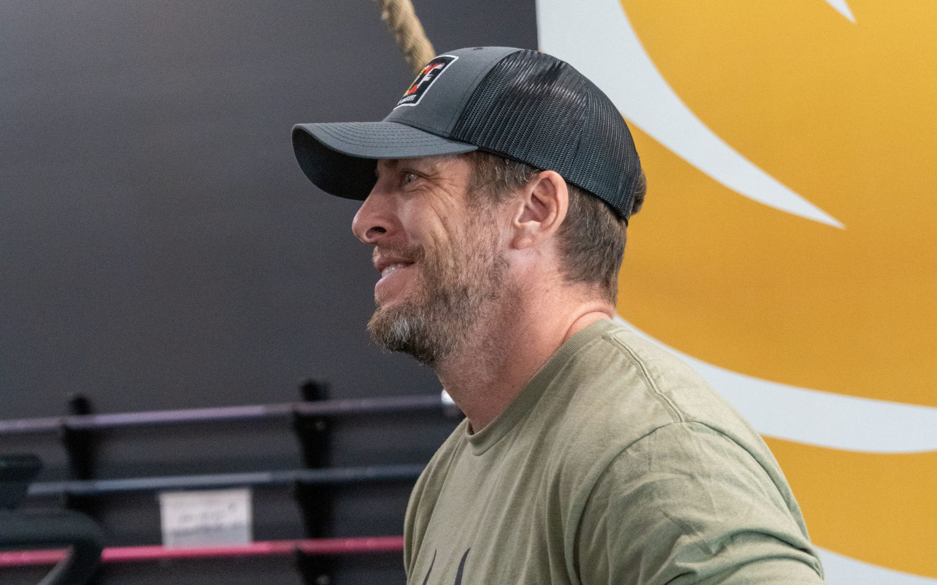 Four people hanging from pull-up bars in a gym, smiling. They are wearing athletic clothing, near weights and equipment.