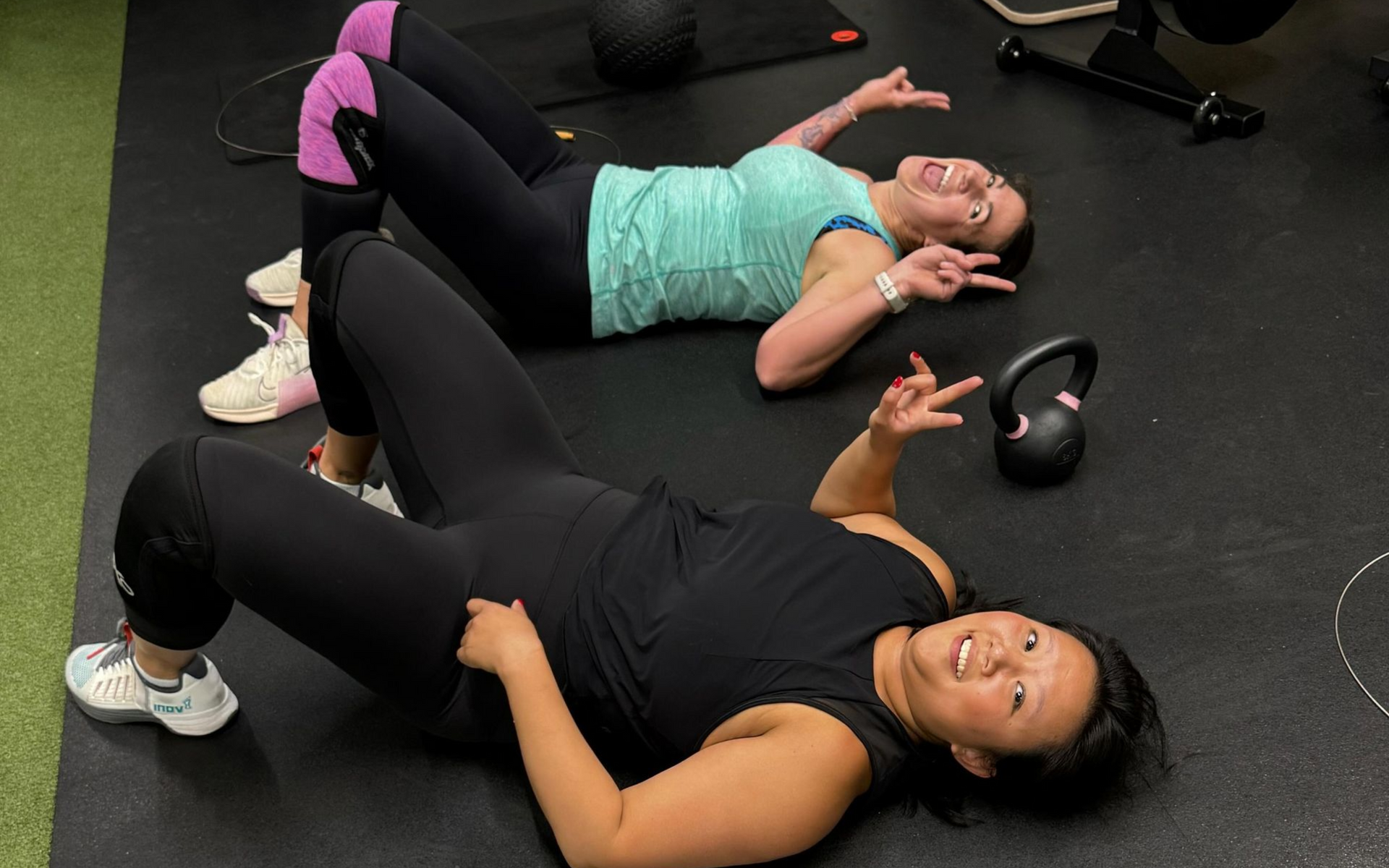 Two people lying on a gym floor in workout clothes, making peace signs, kettlebell nearby.
