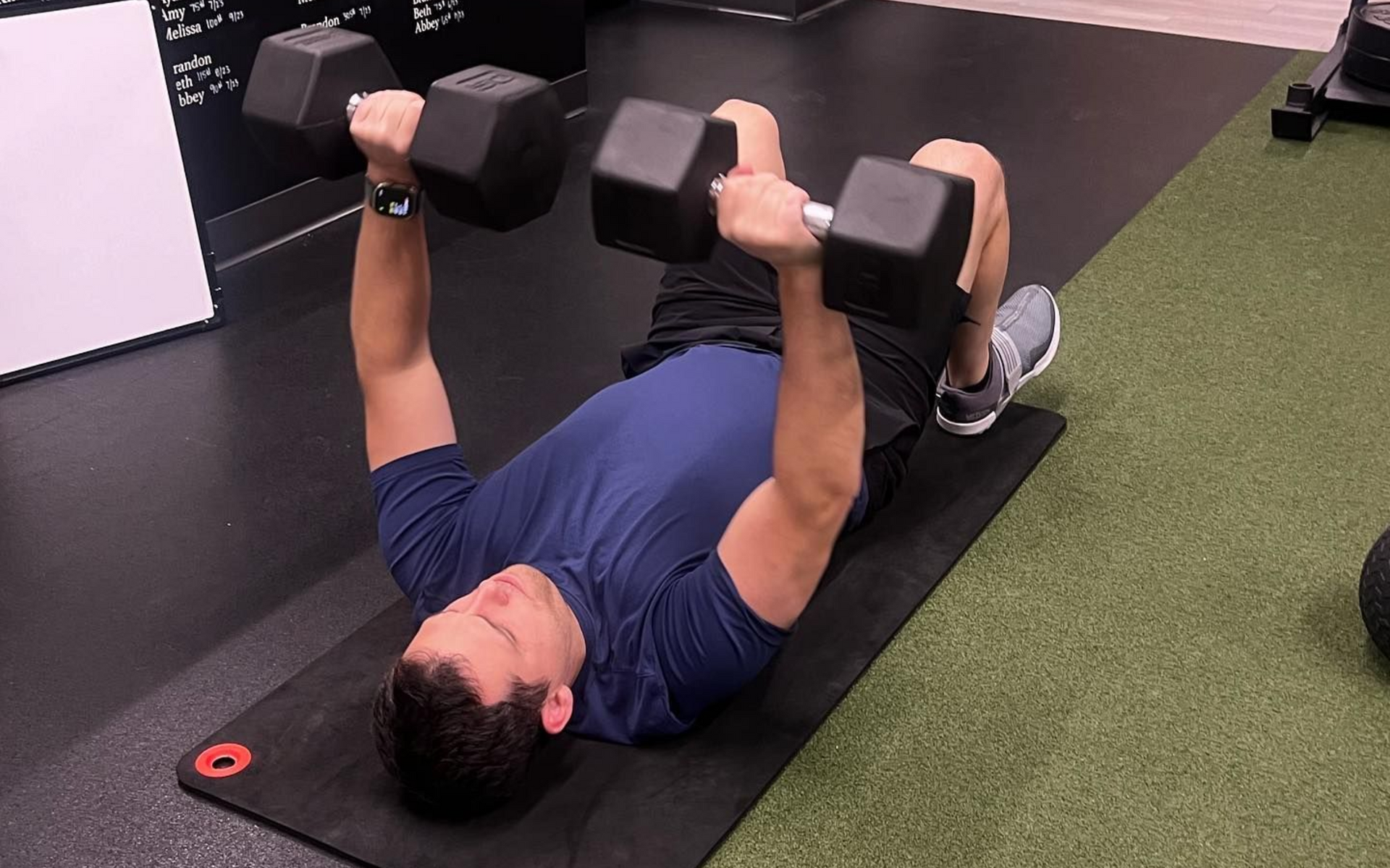 Man on a mat doing dumbbell chest press exercise in a gym.