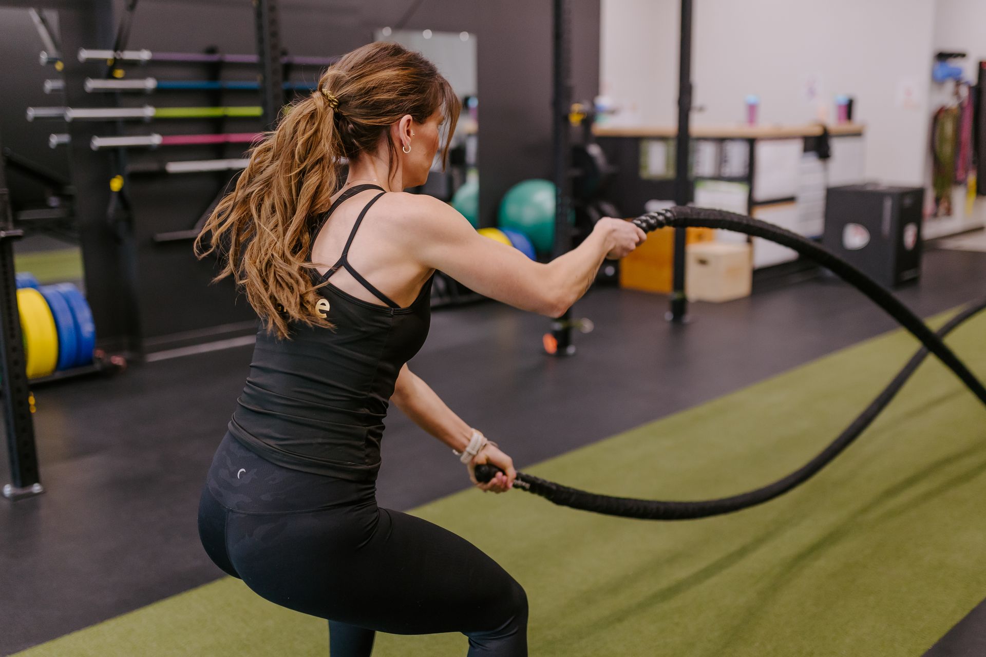 Woman in gym uses battle ropes for a workout on green mat.