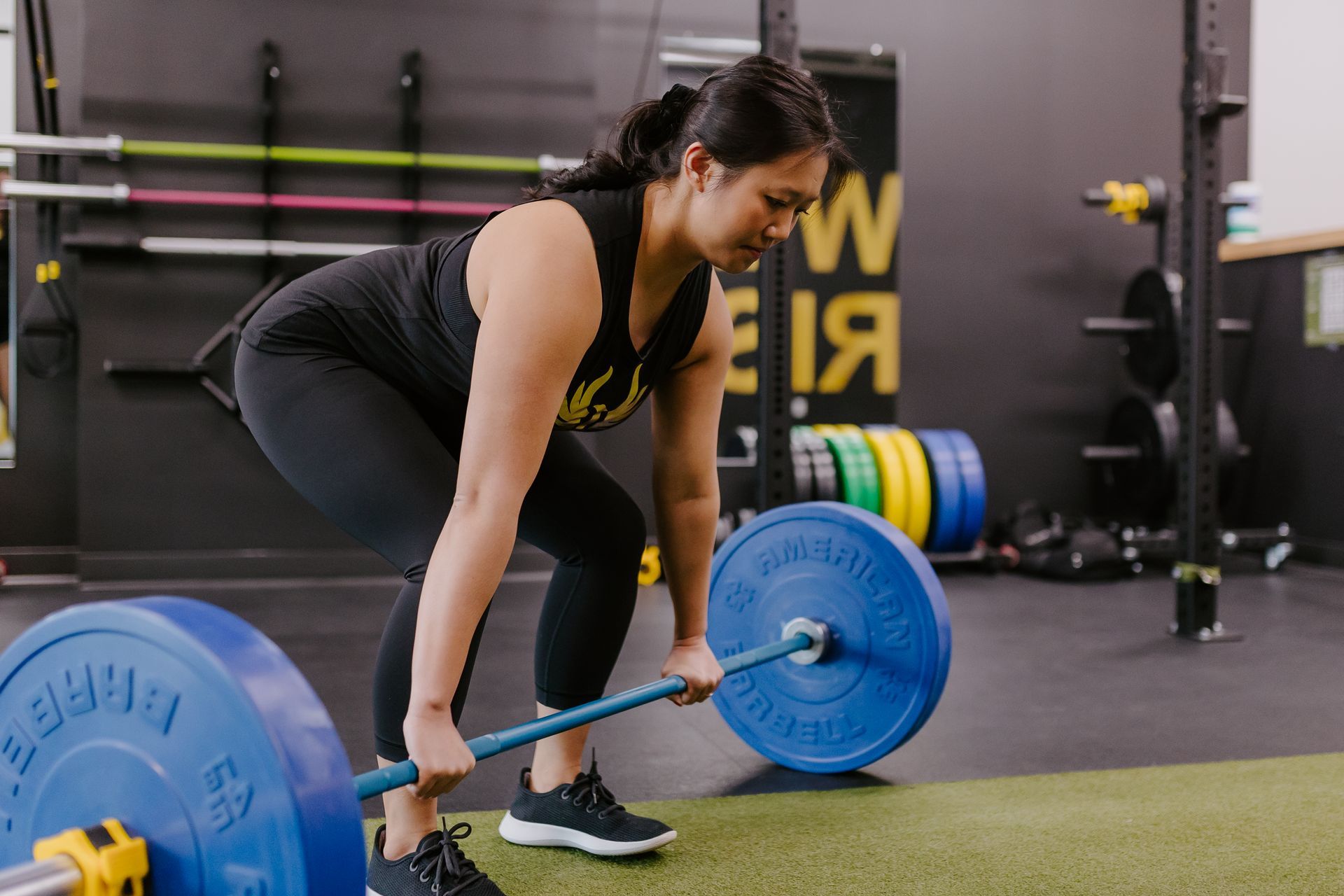Woman in a gym doing a barbell deadlift. She is wearing black activewear, with her hair in a ponytail.