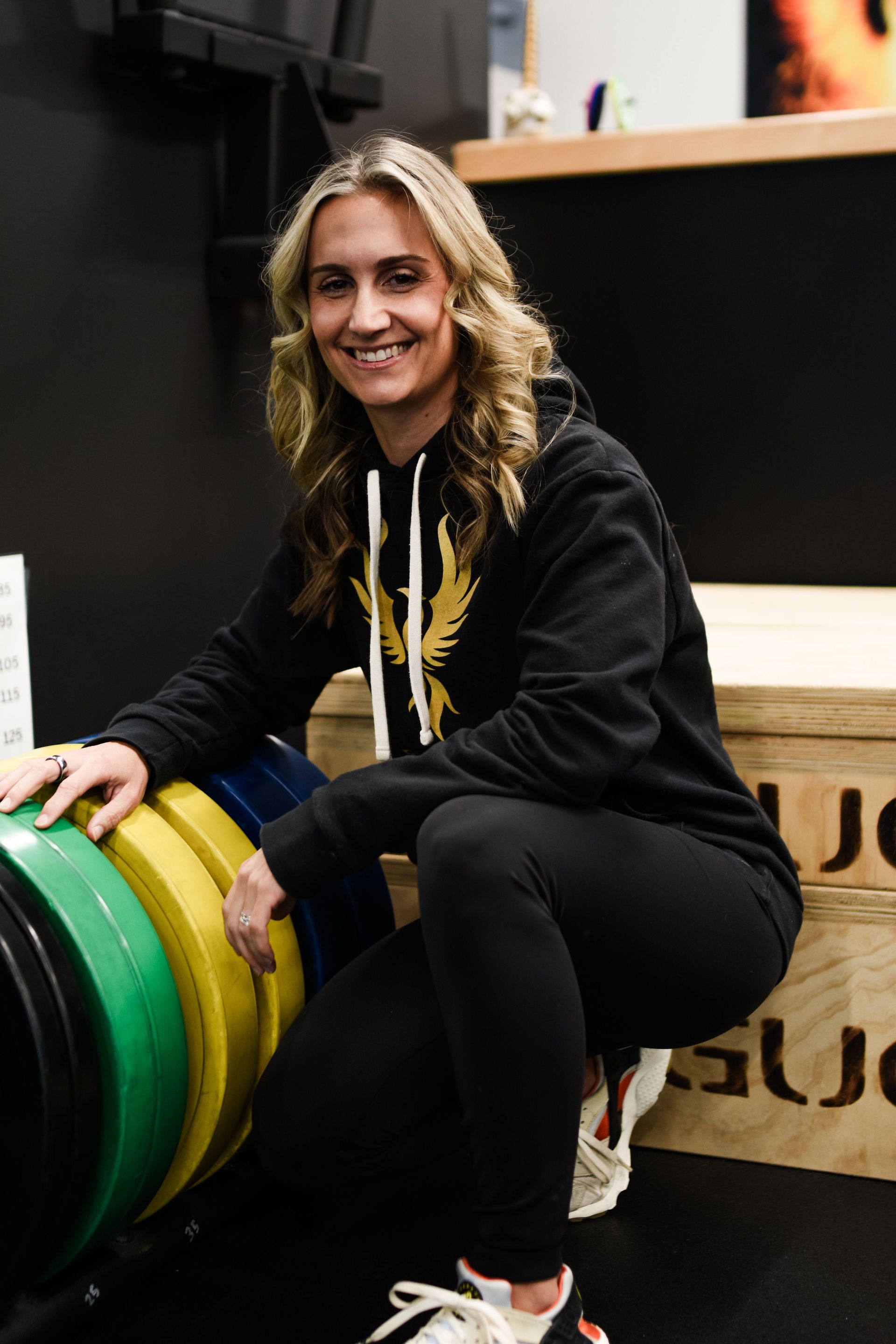 Woman in black hoodie sits near weights, smiling. Indoor gym setting with black background.