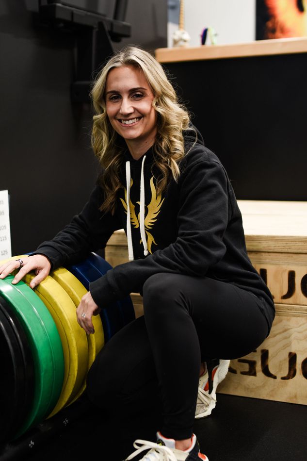 Woman in black hoodie sits near weights, smiling. Indoor gym setting with black background.